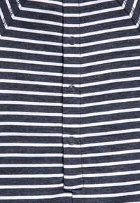 Tkanina v námořnické modři a bílé s proužky, s předním zapínáním na knoflíky, obsahuje pět tmavých knoflíků a výstřih do véčka. Měkká, pletená textura.