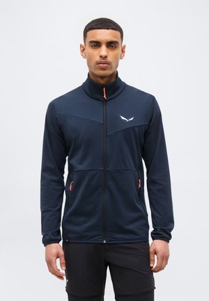 Νεαρός άνδρας που φοράει μπλε μπουφάν με φερμουάρ σε πορτοκαλί χρώμα και μαύρο παντελόνι, στέκεται μπροστά σε ένα απλό φωτεινό φόντο.