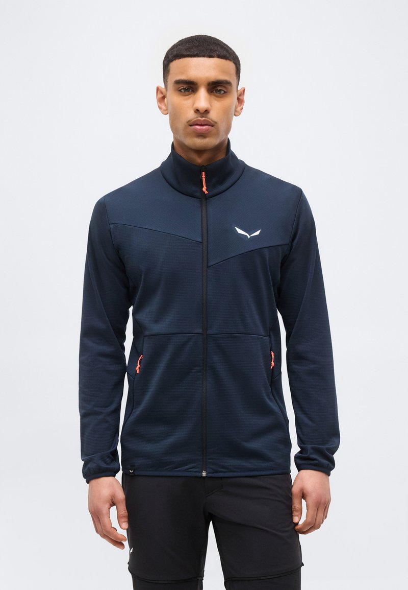 Νεαρός άνδρας που φοράει μπλε μπουφάν με φερμουάρ σε πορτοκαλί χρώμα και μαύρο παντελόνι, στέκεται μπροστά σε ένα απλό φωτεινό φόντο.