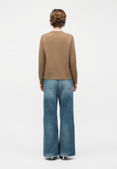 Personne aux cheveux courts et bouclés portant un pull marron et un jean bleu à jambes larges, debout de dos sur un fond blanc uni.