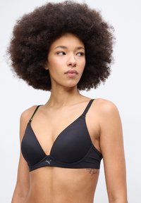 Γυναίκα με μεγάλο σγουρό αφρο μαλλί που φοράει μαύρο σουτιέν Puma, κοιτάζοντας προς τα δεξιά της, με φόντο ένα λευκό, απλό φόντο.