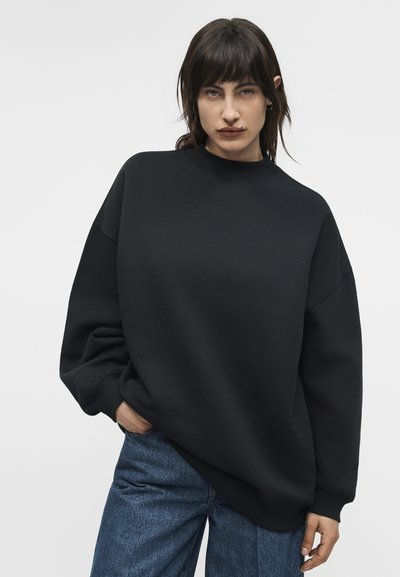 Γυναίκα με σκούρα μαλλιά, ντυμένη με φαρδιά μαύρη φούτερ μπλούζα και μπλε τζιν, στέκεται με το ένα χέρι στην τσέπη, μπροστά σε απλό φωτεινό φόντο.