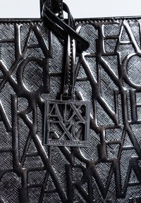 Černá texturovaná kabelka s raženým logem a čtvercovou kovovou štítkem s názvem značky. Hladký povrch s lesklými akcenty.