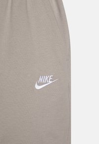 Nike tepláky ve světlé béžové barvě, vyrobené z měkké látky, s úzkým střihem a bílým vyšitým logem na levém stehna.