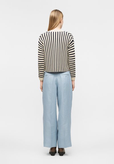 Žena so rovnými blond vlasmi, oblečená v krémovo-čiernom svetri s vertikálnymi pruhmi, svetlomodrých nohaviciach s širokými nohavicami a čiernych topánkach, otočená chrbtom.