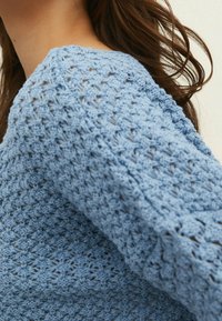 Pull en maille bleu clair avec un motif ouvert et texturé et un col rond, présentant des détails tissés distincts sur le tissu.