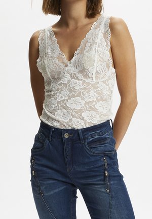 Kvinde iført en hvid blomsterblonde ærmeløs top kombineret med mørkeblå jeans med lynlåslommer foran.