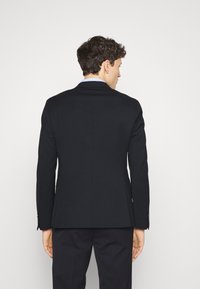 Giacca da completo nera con vestibilità su misura, apertura singola e dettagli di cucitura sottili. Il tessuto appare liscio con una forma strutturata e maniche lunghe.