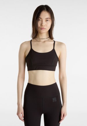 Schwarzer Sport-BH mit dünnen Trägern, glatter Textur und kontrastierenden Nähdetails. Kombiniert mit hochgeschnittenen schwarzen Leggings mit Logo.