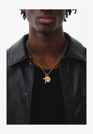 Ung person iført lagdelte guldkæder med kors- og medaljonvedhæng, sort skjorte og sort læderjakke.