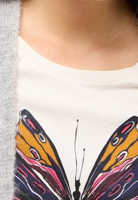 Biała koszulka z dużym, kolorowym motylem w odcieniach pomarańczowego, różowego i niebieskiego, częściowo zakryta miękkim, szarym kardiganem.