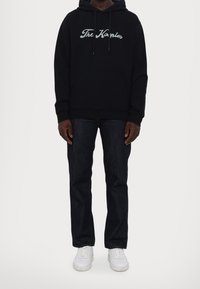 Osoba oblečená v černé mikině s nápisem "The Kooples", tmavých džínách a bílých teniskách, stojící na světlém pozadí.