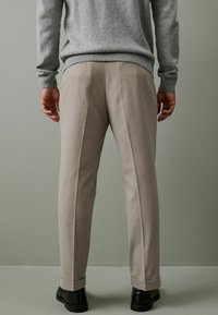 Beige Hosen mit maßgeschneidertem Schnitt und Faltenstepp, kombiniert mit einem grauen Pullover, von hinten betrachtet. Schwarze Schuhe vervollständigen das Outfit.