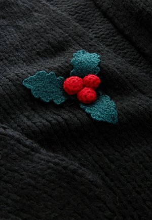 Crni teksturirani materijal sadrži dekorativnu aplikaciju zelenih listova i tri crvene pom-pom bobice u svečanom aranžmanu.