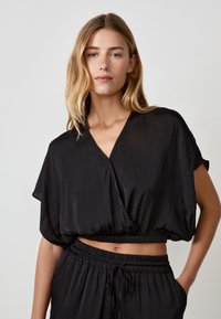 Blusa nera corta con scollatura a V, maniche corte e vita elasticizzata, realizzata in un tessuto leggero e liscio. Abbinata a pantaloni con vita elastica.
