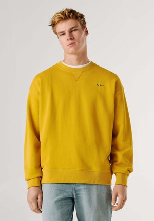 Joven con cabello corto rubio que lleva una sudadera amarilla mostaza y jeans azul claro, de pie contra un fondo blanco liso.