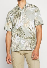 Mies, joka pukeutuu lyhythihaseen napitettuun paitaan, jossa on beigen, valkoisen ja vihreän trooppisen lehden kuvio, yhdessä vaaleanbeigeiden cargo-housujen kanssa.