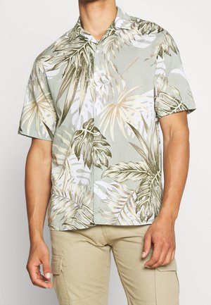 Man in een kortemouw drukknopen shirt met een lichtbeige, witte en groene tropische bladerenprint, gecombineerd met lichtbeige cargobroeken.