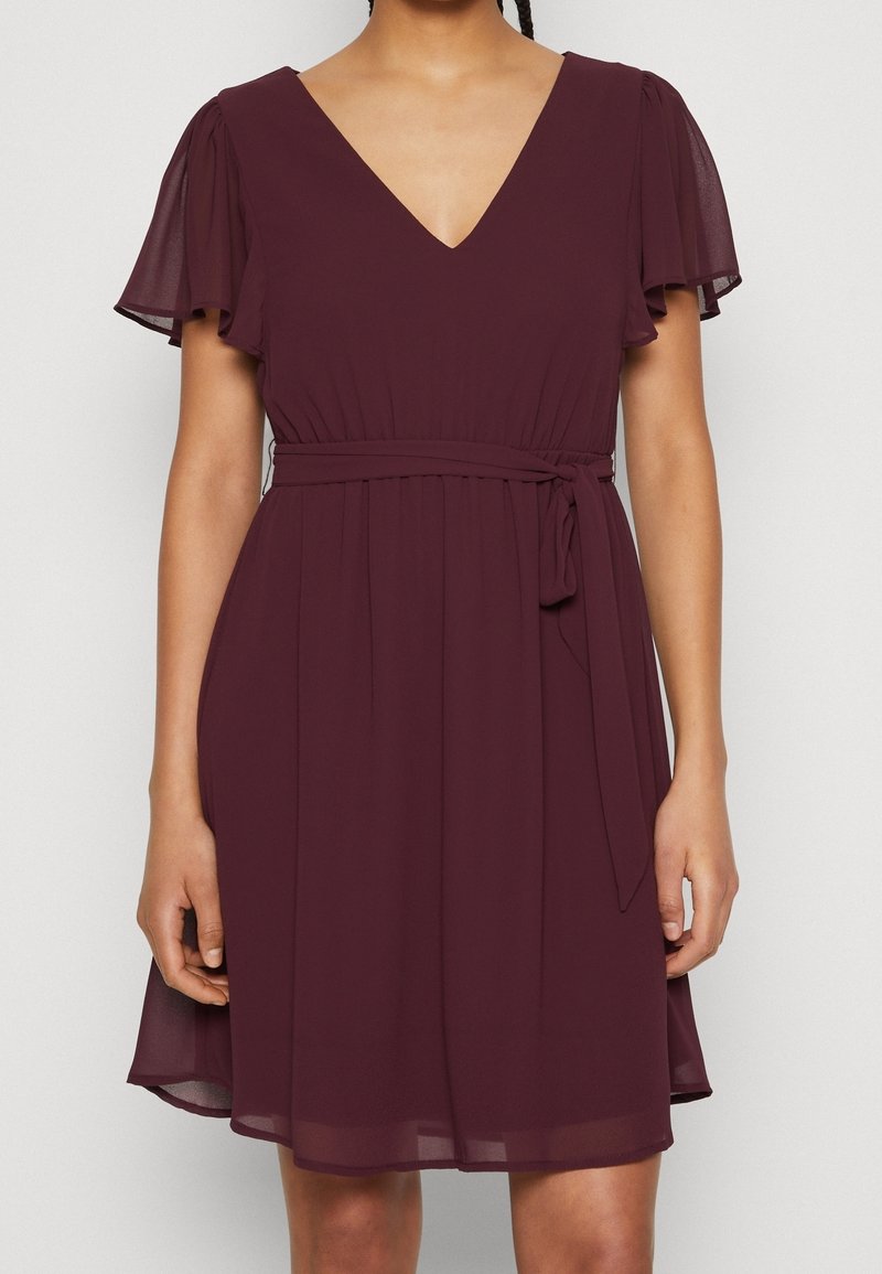 Femme portant une robe prune à manches courtes, col en V, longueur genou, avec ceinture à nouer à la taille et manches volantées.
