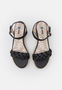 Černé sandály s pleteným popruhem, s přezkou kolem kotníku a tkanou podrážkou v espadrilovém stylu. Vnitřní podrážka má vzor listů na béžovém pozadí.