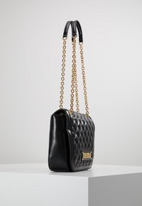 Vadderad svart läderväska med detaljer i guldkedja och en strukturerad form. Har en logotavla och slät yta.