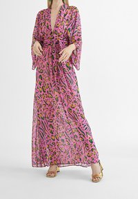 Maxiklänning i genomskinlig rosa tyg med ett djärvt djurmönster i grönt och gult. Har vida ärmar och en knuten midja. Sandalerna är gyllene.