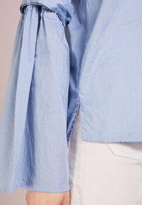 Chemise bleu clair à manches longues avec manches évasées froncées, présentant une coupe décontractée et des fentes latérales pour plus de liberté de mouvement et de confort.