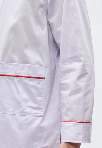 Λεπτομέρεια λευκού μπουφάν με μακριά μανίκια και τσέπη, με κόκκινη διακοσμητική ραφή κατά μήκος της κορυφής της τσέπης και της μανσέτας, πάνω σε απλό φόντο.