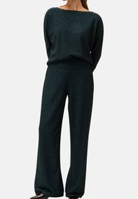 Femme portant un haut texturé à manches longues vert foncé et un pantalon à jambes larges avec des chaussures pointues noires, debout devant un fond blanc uni.