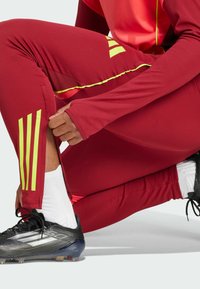 Rote Sporthosen aus flexiblem Material mit gelben Akzenten und drei Streifen. Ein schwarzer Fußballschuh ist sichtbar, kombiniert mit weißen Socken.