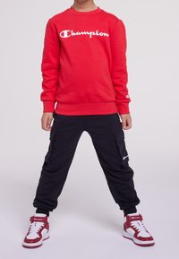Felpa rossa con logo bianco "Champion", pantaloni cargo neri e sneakers alte rosse e bianche. Abbigliamento sportivo comodo e informale.