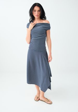 Vrouw met donker haar die een blauw-wit gestreepte off-shoulder jurk en gouden teenslippers draagt, staand op een witte achtergrond.
