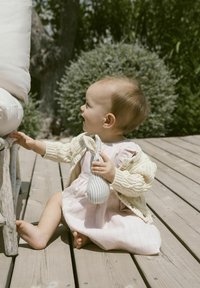 Strickcreme-Cardigan, rosa Kleid mit vertikalen Streifen, barfuß sitzendes Kind auf einer Holzterrasse, das ein gestreiftes Stoffspielzeug hält und lächelt.