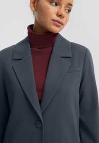 Blazer grigio con una texture liscia, dotato di colletto a scollo, chiusura con un bottone singolo e taschino sul petto, indossato sopra un dolcevita bordeaux.