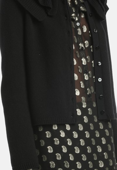 Schwarze, gestrickte Strickjacke mit einem gerippten Kragen und Knöpfen, über einer transparenten Bluse mit einem silbernen Paisleymuster auf schwarzem Hintergrund getragen.