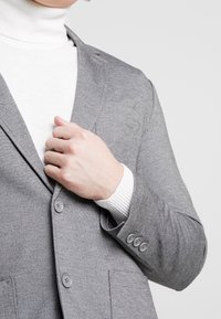 Blazer grigio con finitura testurizzata, dotato di due bottoni e vestibilità sartoriale, indossato sopra un dolcevita bianco. Dettaglio dei polsini notevole visibile.