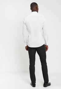 Weißes Langarmhemd mit klassischem Kragen, kombiniert mit dunklen Hosen und schwarzen Schuhen; glatte Textur und taillierte Passform.