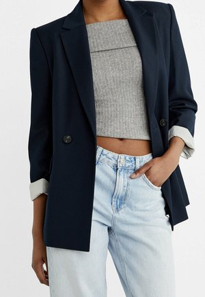 Personne portant un blazer bleu marine aux manches retroussées, un crop top gris côtelé et un jean taille haute bleu clair, une main dans la poche.