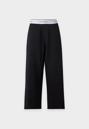 Jeune homme debout portant un t-shirt rouge Dsquared2, un pantalon large noir et des chaussures bordeaux sur un fond blanc uni.