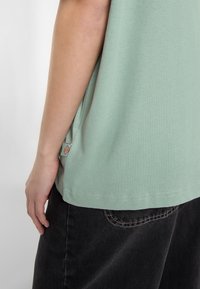 T-shirt en coton vert clair avec une coupe décontractée, un ourlet incurvé et une petite étiquette logo sur le côté. Accompagné de pantalons gris foncé.