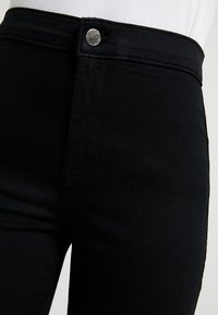 Svarta tajta byxor med silvrig knäppning i midjan, bärs tillsammans med en vit topp.