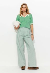 Haut en tricot vert avec col blanc et rayures, associé à un pantalon large à rayures vert clair. Le modèle tient un objet blanc, portant des chaussures à imprimé animal.