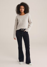 Donna con capelli ricci che indossa un maglione beige con bottoni, pantaloni neri con cintura e sneakers bianche su uno sfondo neutro.