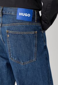 Fittede blå denimjeans med fem-lomme design, prydet med gule syninger og et markant blåt mærke med hvid skrift i taljen.