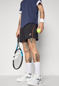 Tennise kostüüm, mis sisaldab tumesinist pluusi valgete detailidega, musti lühikesi püksid oranži äärisega, valget jalatseid ning tennisereketi ja palli hoidev inimene.