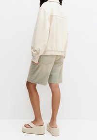 Giacca in cotone beige con orlo elasticizzato, abbinata a pantaloni corti in denim verde chiaro. I sandali con platform sono testurizzati e di un colore neutro chiaro.