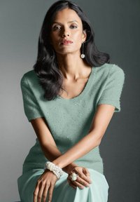 Lichtgroene trui met korte mouwen en een ronde halslijn, met textuur details. Het model draagt parelsieraden en zit met gekruiste armen.