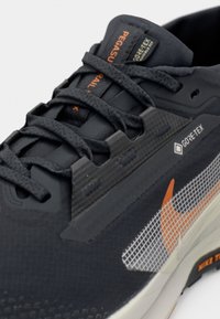 Schwarzer wasserfester Sneaker mit strukturiertem Stoff, grauen Akzenten, orangefarbenem Logo und Gittermuster-Details an der Seite. Verfügt über eine gepolsterte Zunge und Schnürsenkel.