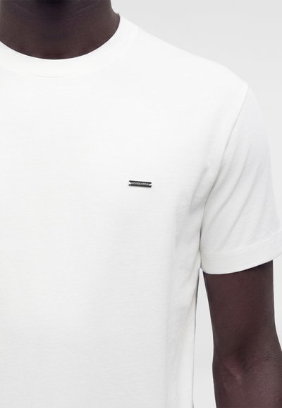 Personne à la peau foncée portant un t-shirt blanc uni à manches courtes avec un petit logo rectangulaire noir et blanc sur la poitrine gauche.