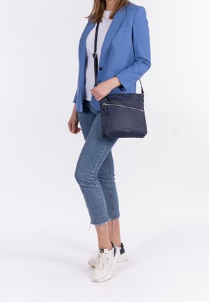 Donna che indossa un blazer blu, camicia bianca, jeans corti, sneakers bianche, con una borsa a tracolla blu navy con una tasca frontale con cerniera.